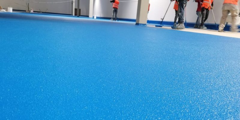 Trabajadores con chaleco naranja enfocados en los acabados de un piso de uretano de colr azul