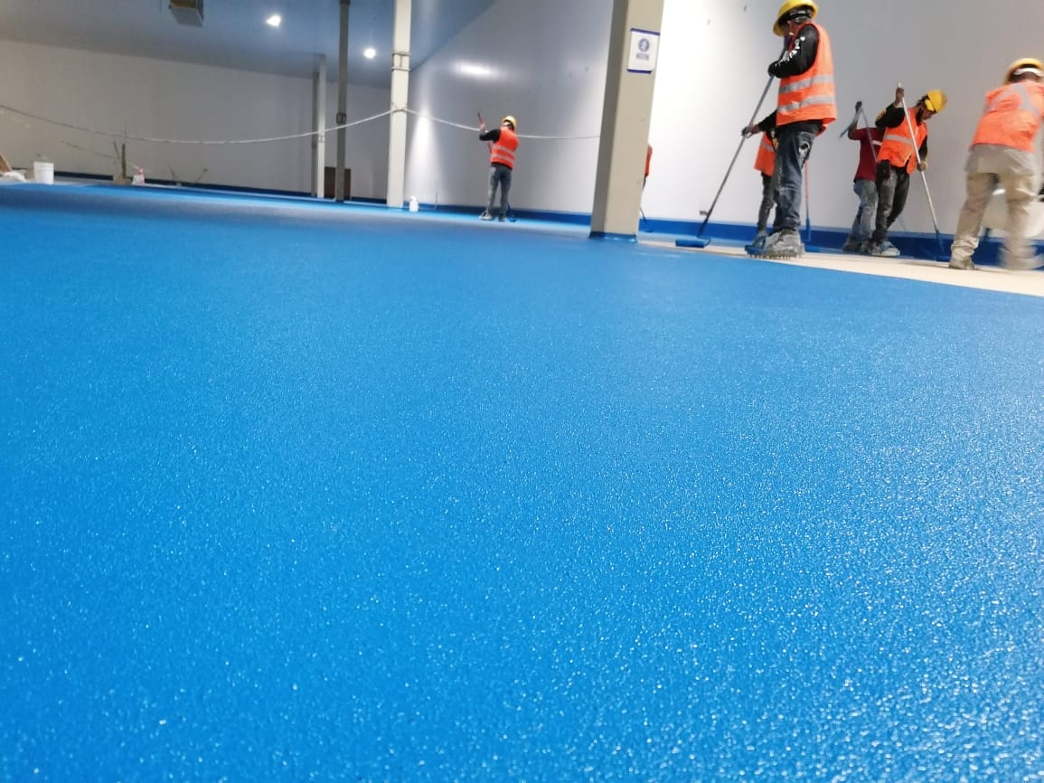 Trabajadores con chaleco naranja enfocados en los acabados de un piso de uretano de colr azul
