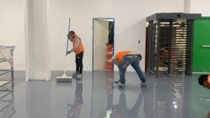 Trabajadores aplicando recubrimiento epóxico en un almacén en construcción, usando rodillos y herramientas, con estanterías vacías y conos de seguridad en el fondo.
