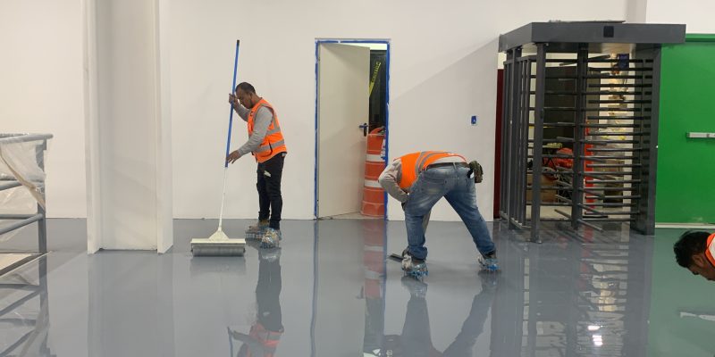 Trabajadores aplicando recubrimiento epóxico en un almacén en construcción, usando rodillos y herramientas, con estanterías vacías y conos de seguridad en el fondo.